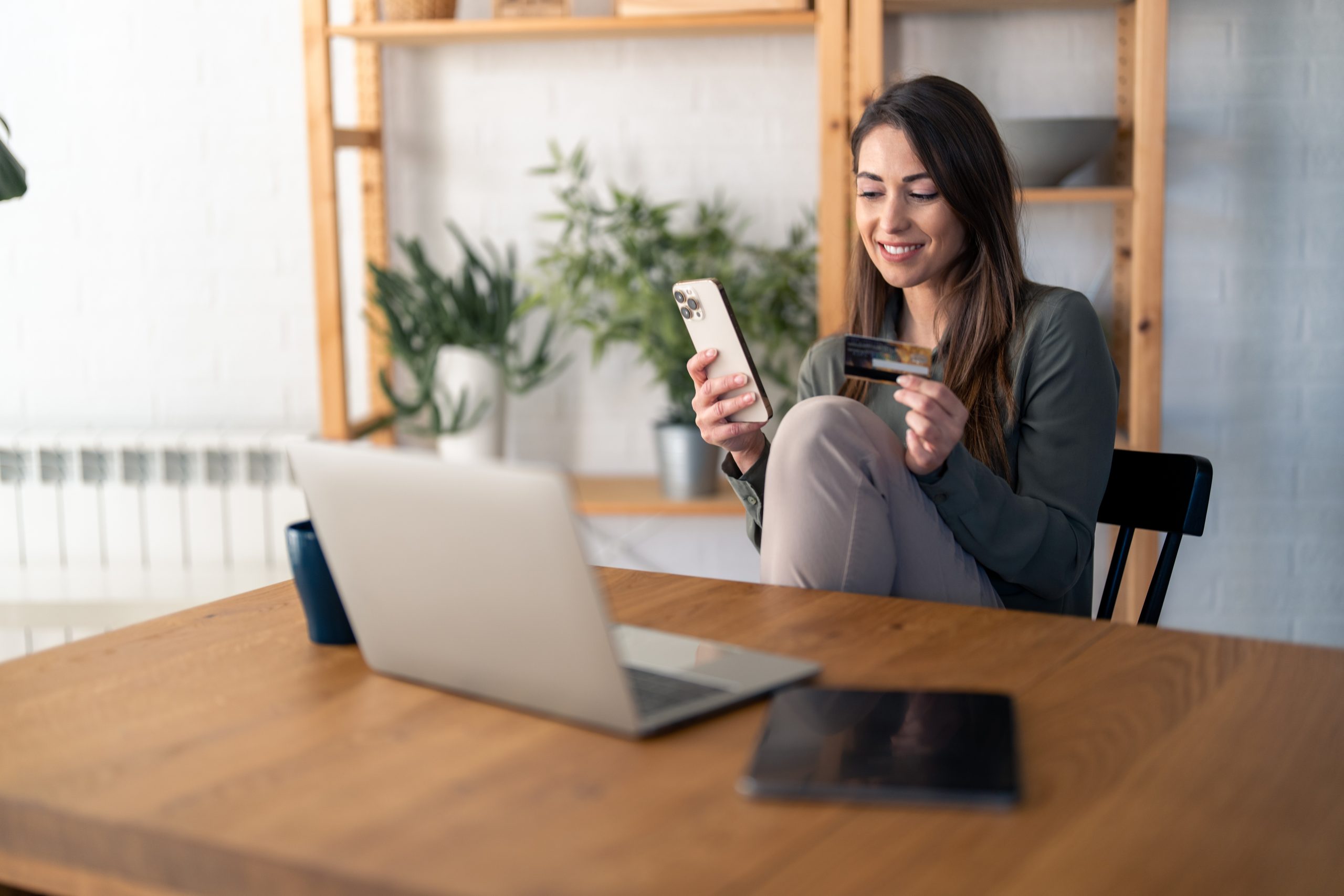 Happy young woman, satisfied online buyer holding credit card, using e-banking app on smart mobile phone making convenient financial e-commerce payment digital transaction while sitting at home.